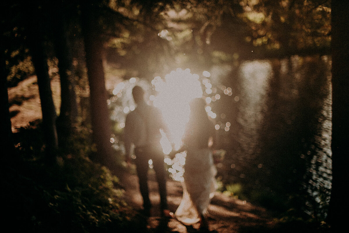 Bride and groom walking away from the camera down a path toward a lake. The image is blurred and the sunlight is reflecting off the lake