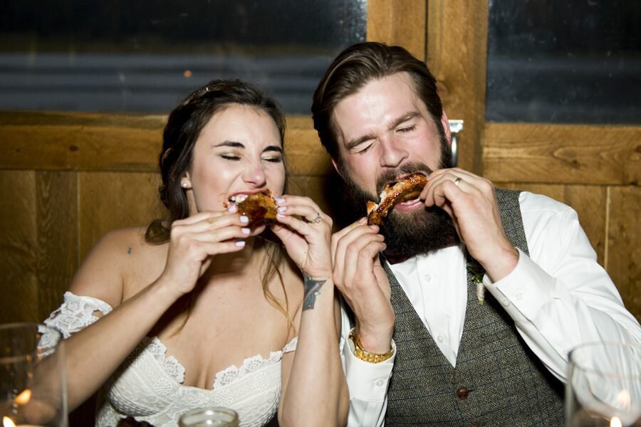 A bride and groom pose while biting into chicken wings at their wedding reception.