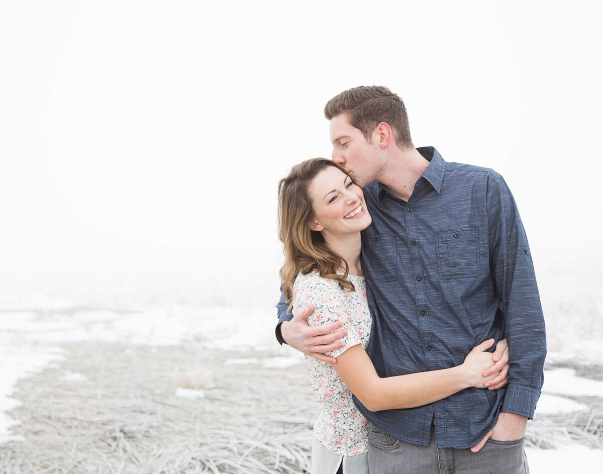 A man in a blue shirt kissing his fiance in a frosted wonderland by Utah Lake