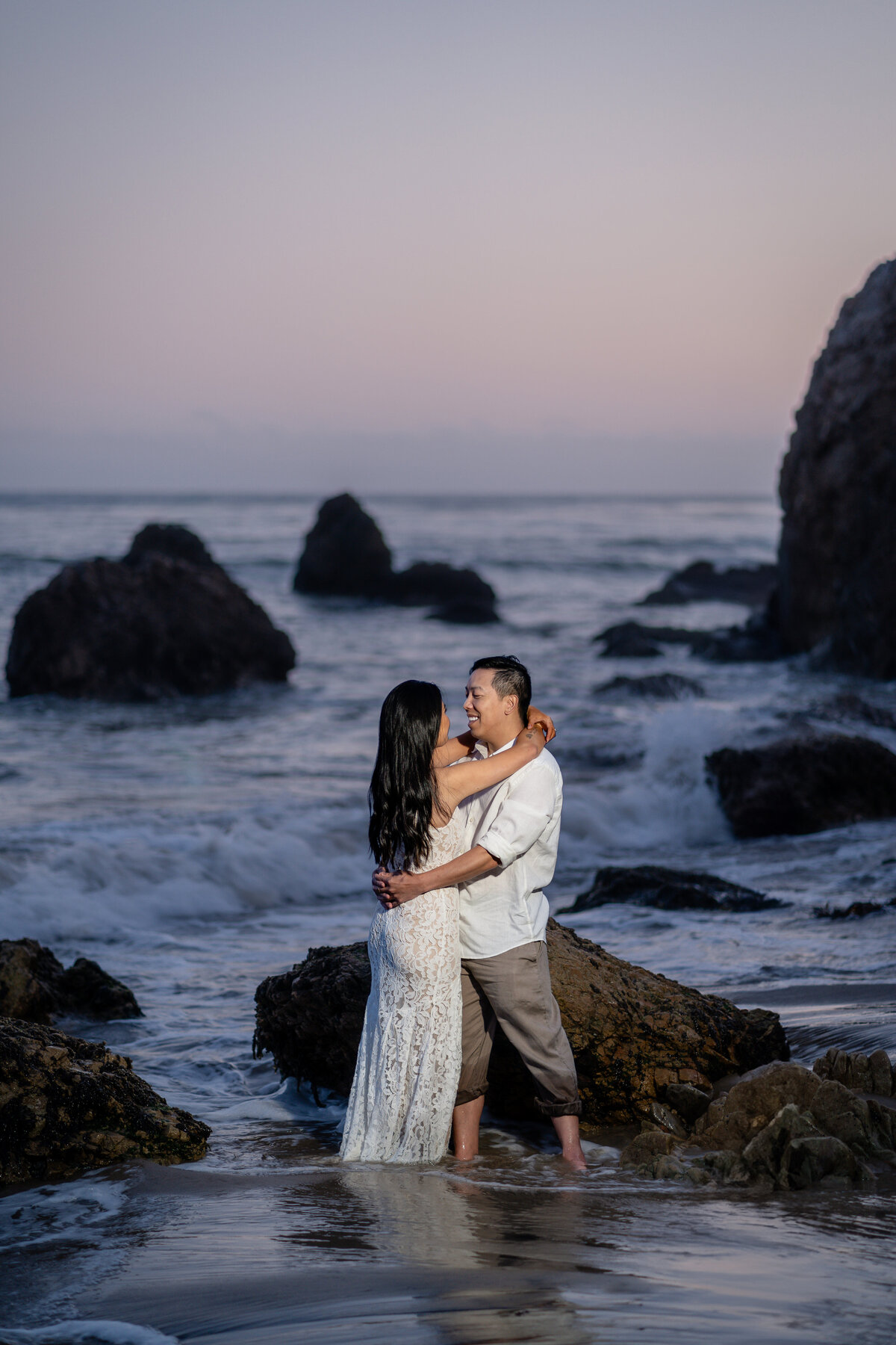 Asian couple eloping at El Matador State Beach in Malibu during low-tide sunset and embracing with rocky cliff beach and ocean waves.