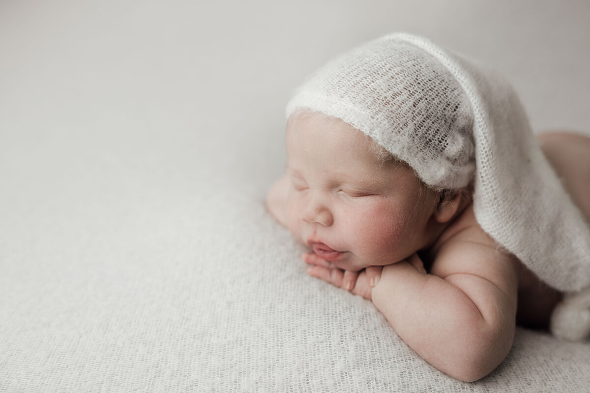 Studio newborn photography - Side profile image of baby sleeping  with his hands resting under his chin. Baby is wearing a long knit hat trailing over his shoulder