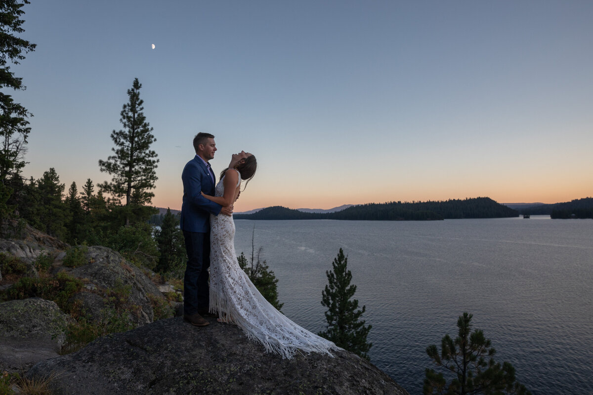 A groom and bride stand facing each other as the bride throws her head back in laughter.