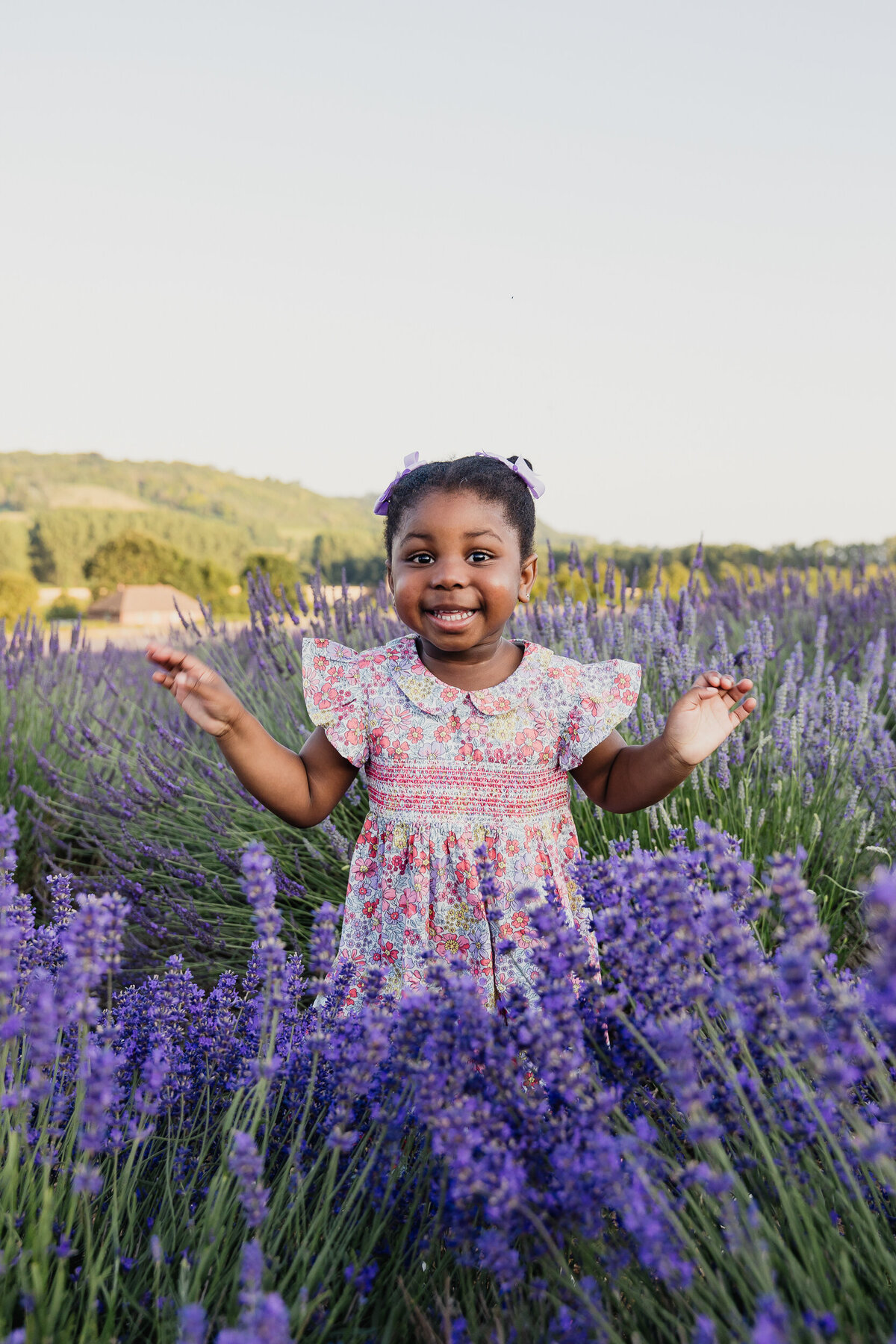 A joyful young girl with a floral dress and bow in her hair stands in a field of lavender, smiling and spreading her arms, with hills in the background.