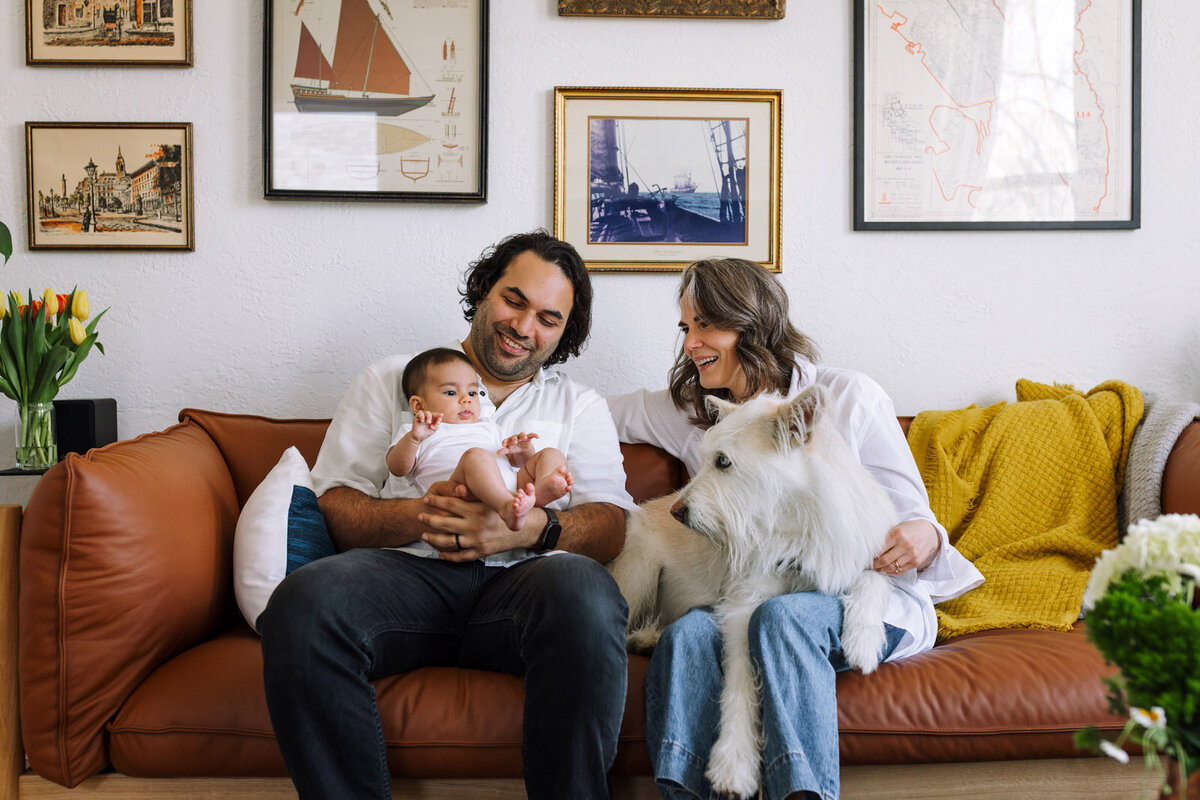 dad holding baby on couch and mom sitting with white dog in her look with everyone looking at baby