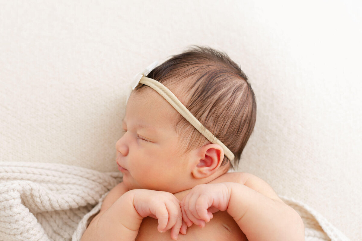 Baby is sleeping on a cozy beige blanket, she has her head turned to one side showing us a profile view of her and she has her arms curled up under her chin.