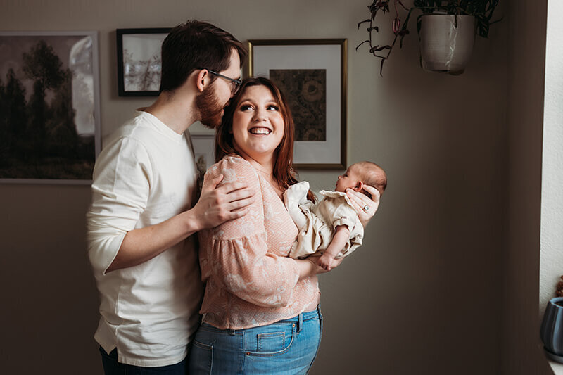 husband stands behind wife and kisses her head while she holds a newborn baby girl on her chest