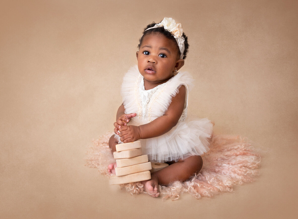 Baby girl sitting for a 6-month milestone photoshoot.Baby is wearing a white tulle outfit with wooden blocks stacked between her legs. She is looking at the camera.