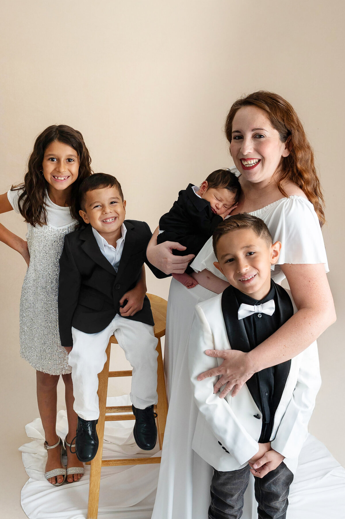 Woman holding a newborn baby and three children wearing coordinating black and white outfits, standing close together and hugging affectionately.