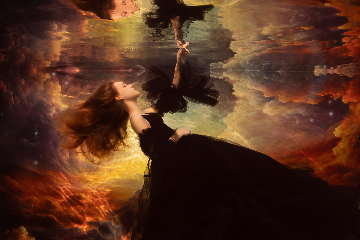 A young woman in a flowing black gown floating underwater, reaching toward her own reflection against a dramatic cloudscape of golden and fiery tones, captured by a DFW underwater photographer.
