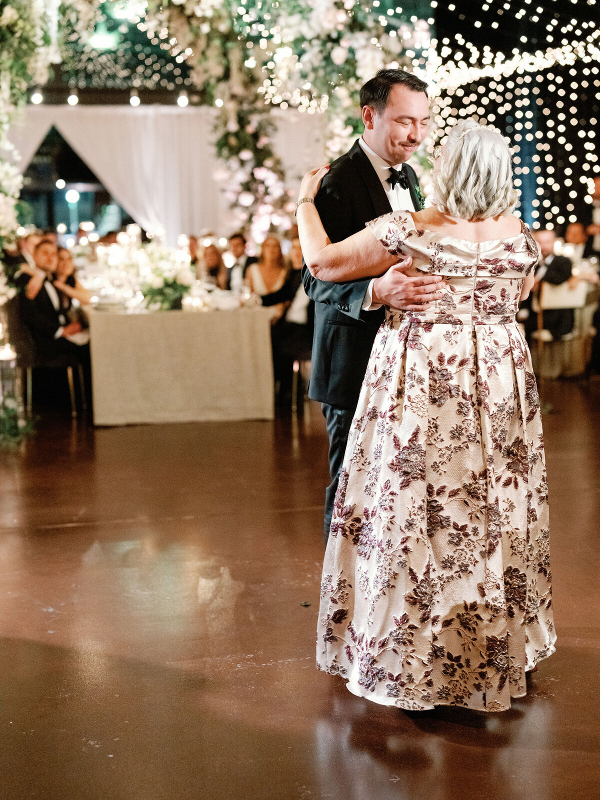A groom shares a heartfelt dance with his mother at the wedding reception, surrounded by guests and a beautifully decorated venue.