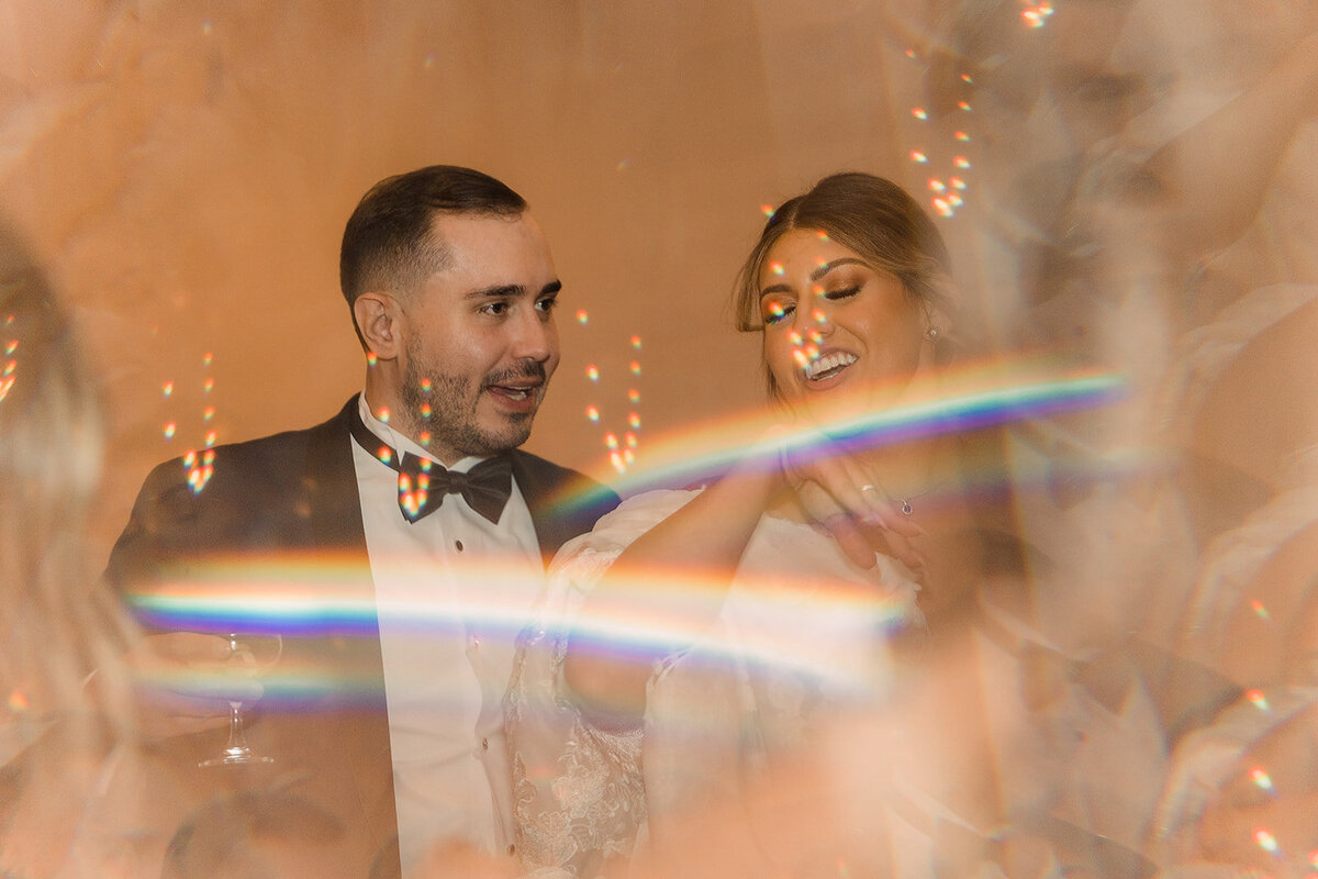 The bride and groom are framed through a prism effect, creating a dreamy, rainbow-like overlay as they share a joyful moment on the dance floor.