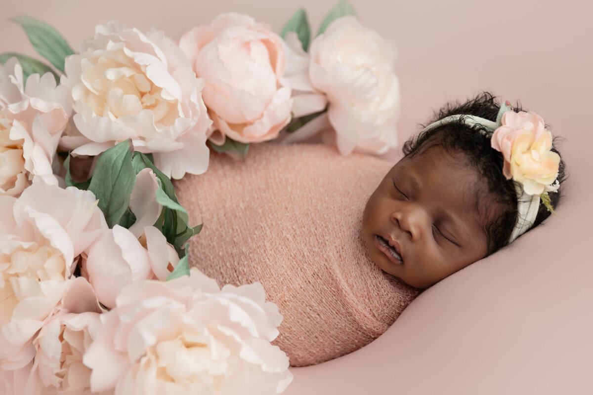 A newborn baby sleeps covered in flowers in a pink swaddle in a studio