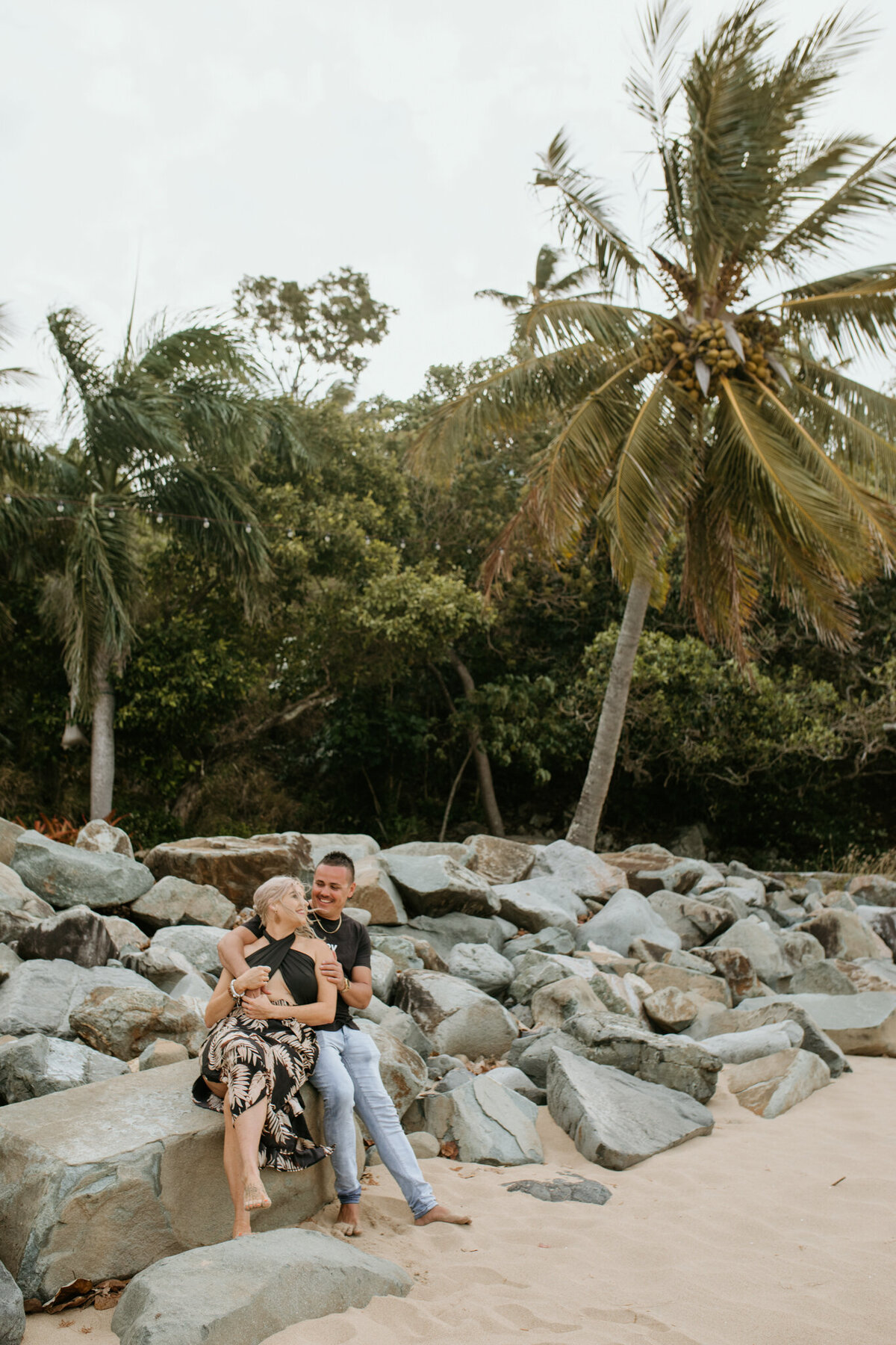 A cute cuddle shared between a couple on a beach, looking at each surrounded by  palm trees.