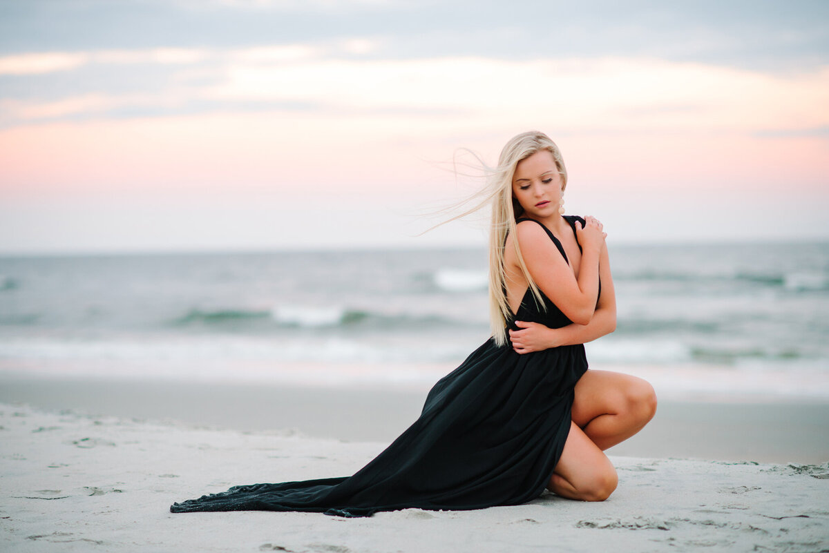 Girl in black dress seated in sand at the edge of the water during a Charleston senior portrait session

