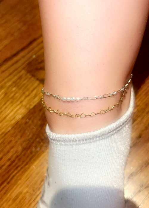 A close-up of dainty permanent anklets in gold and silver with heart chain and pearl accents.