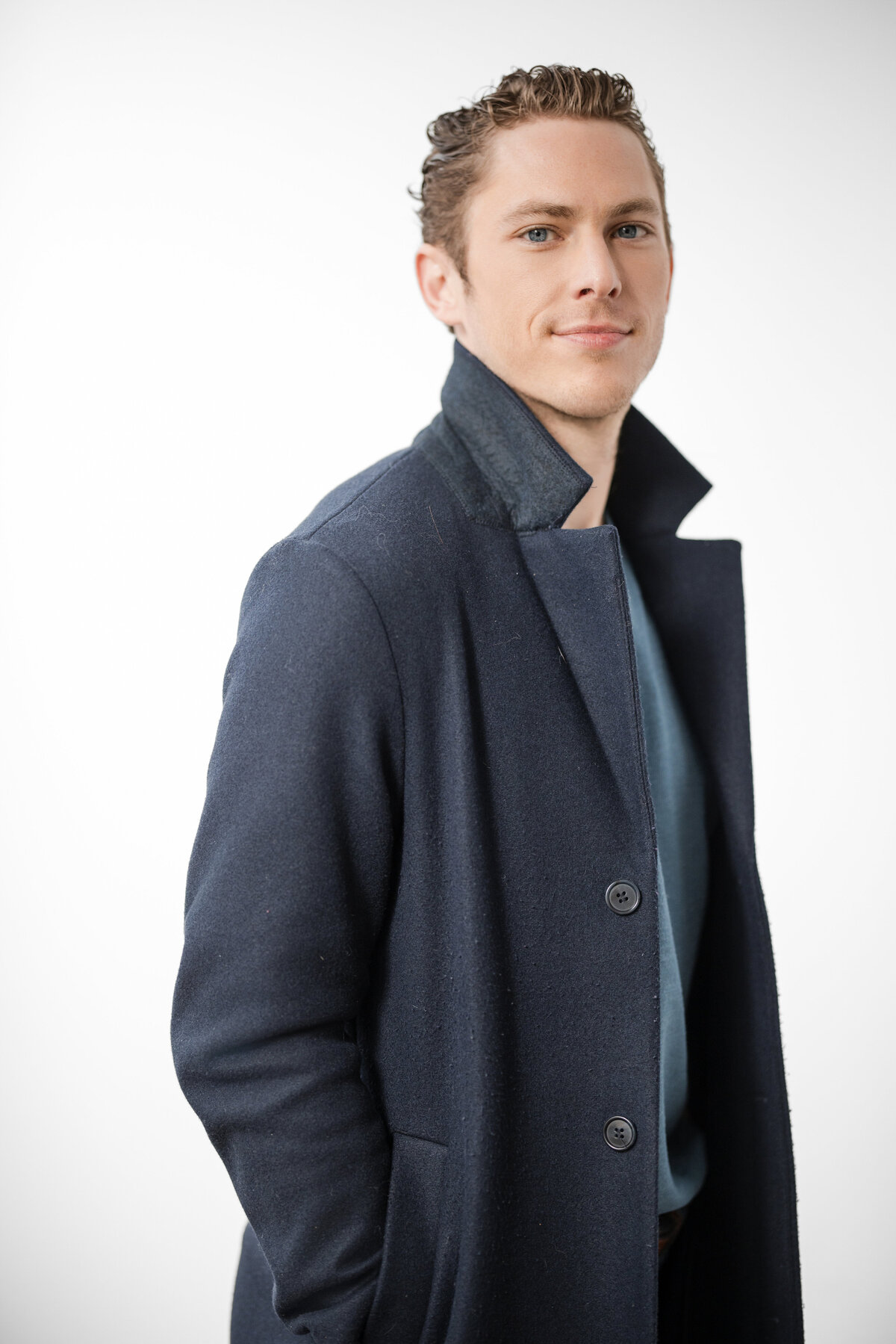 Person standing against a bright white background, wearing a navy coat with the collar popped and a confident expression. Their hands are in their pockets, and they are looking slightly off-camera. The portrait is modern and stylish with soft, natural lighting.
