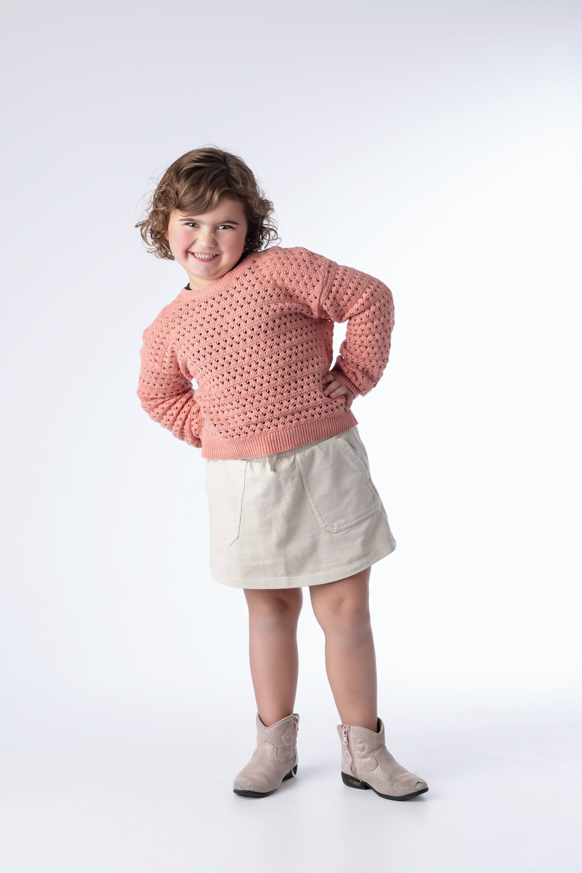A little girl in a pink sweater and white skirt stands on a white background