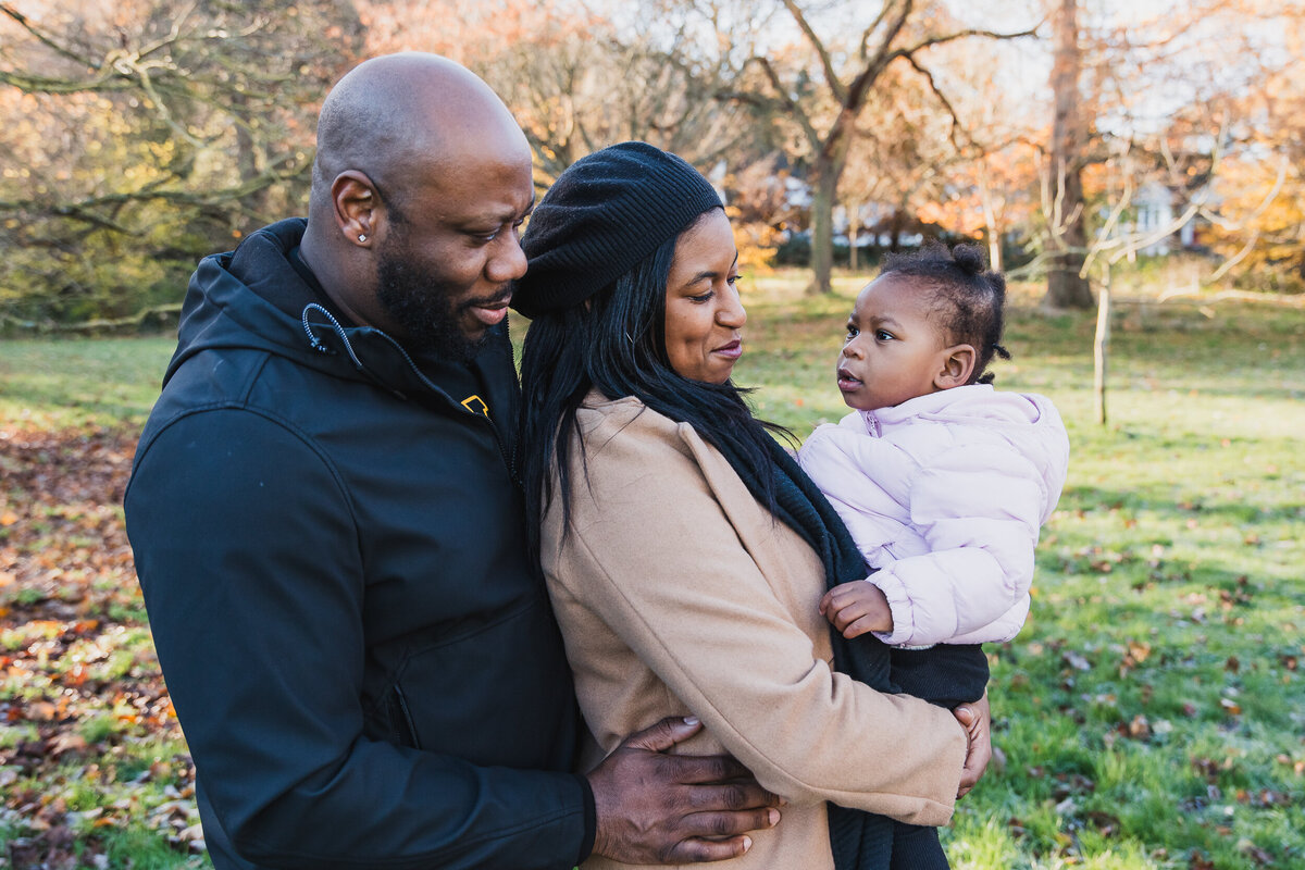 A couple holding their toddler in a park during autumn, smiling and enjoying a sunny day.