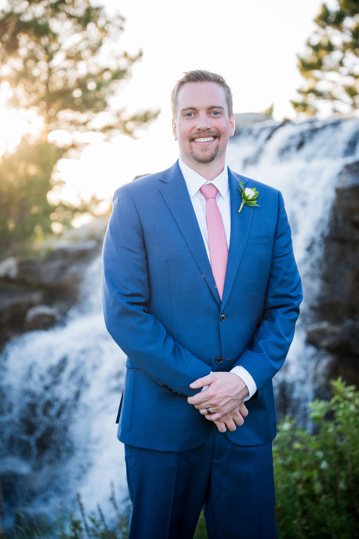 A groom stands with his hands crossed in front, smiling at the camera with a golf course waterfall in the background.