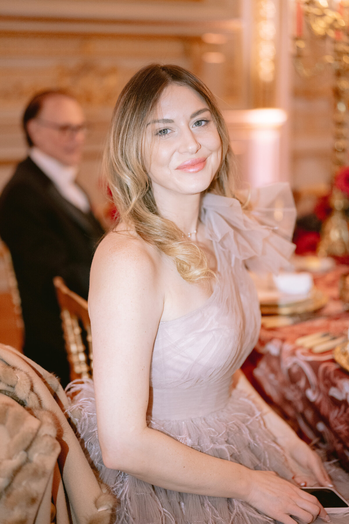 The stunning photo was captured by the talented photographer Matteo Coltro at the luxurious Shangri La Hotel & Ritz Paris Hotel in Paris, France. Zen Film Works also played a crucial role as videographers in capturing the moments on video. In the image, a woman with light brown hair styled in loose curls looks radiant as she sits at a table adorned with ornate red and gold decorations. She is dressed in a gorgeous light pink, one-shoulder dress embellished with delicate tulle and feathers, accentuating her elegant smile directed towards the camera. Amidst the opulent setting, a blurred figure of a man in a suit adds a sense of movement and intrigue to the scene.