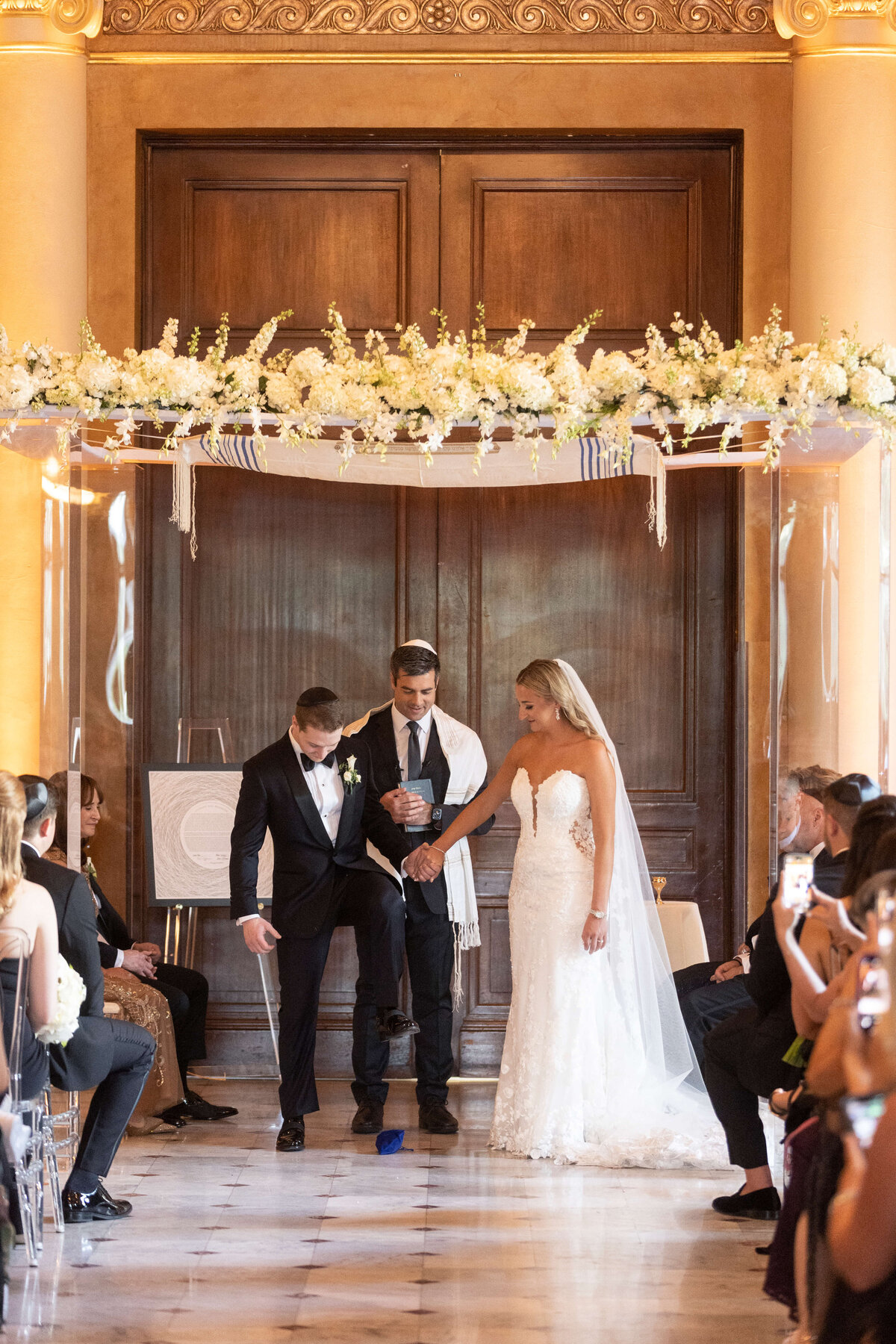 A bride in a white gown and veil holds hands with a groom in a tuxedo breaking a glass under an ornate floral chuppah during a wedding ceremony. Guests are seated on either side, watching the couple.