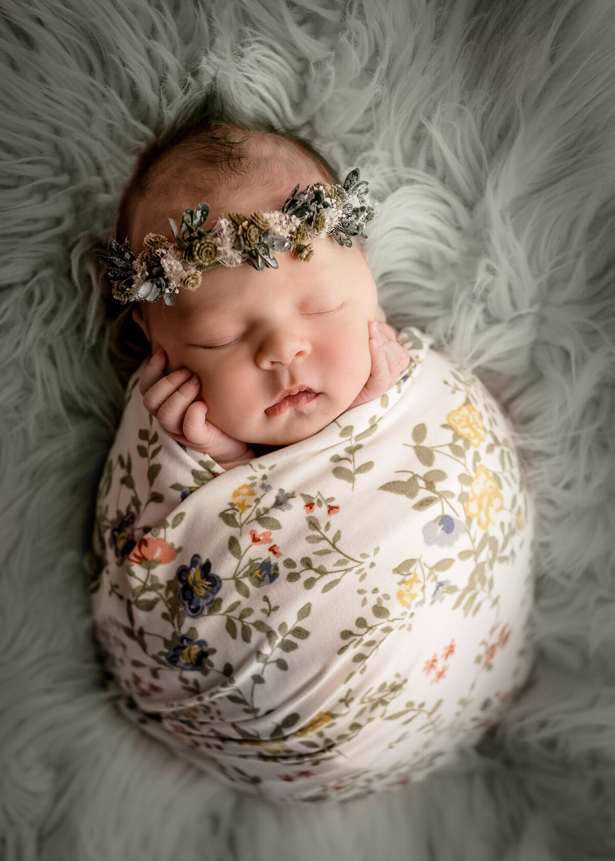 A newborn baby girl sleeps with hands on her cheeks in a floral swaddle for a Des Moines Newborn Photographer