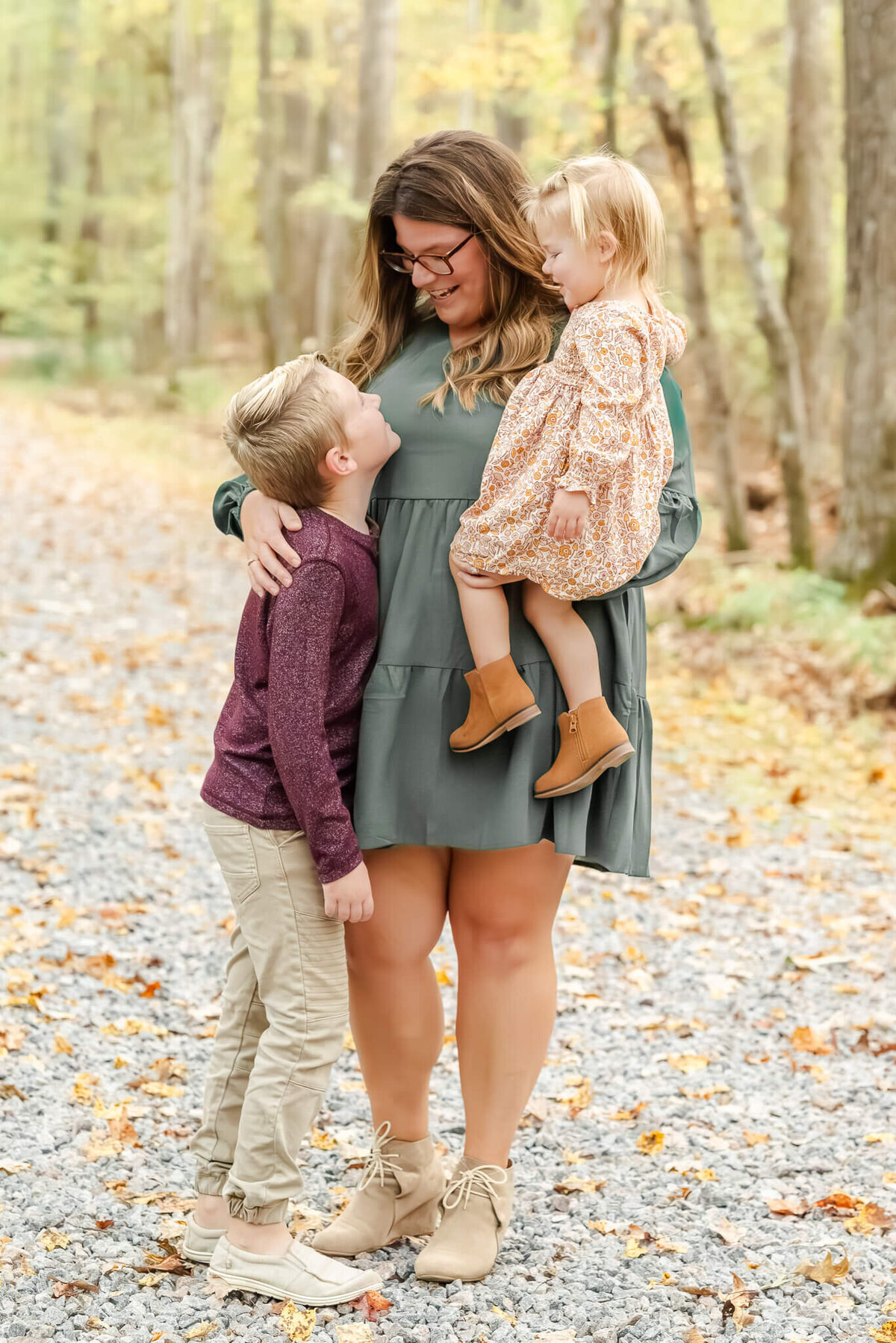 A mom wearing a teal dress holds her young daughter on her hip while looking lovingly down at her son.