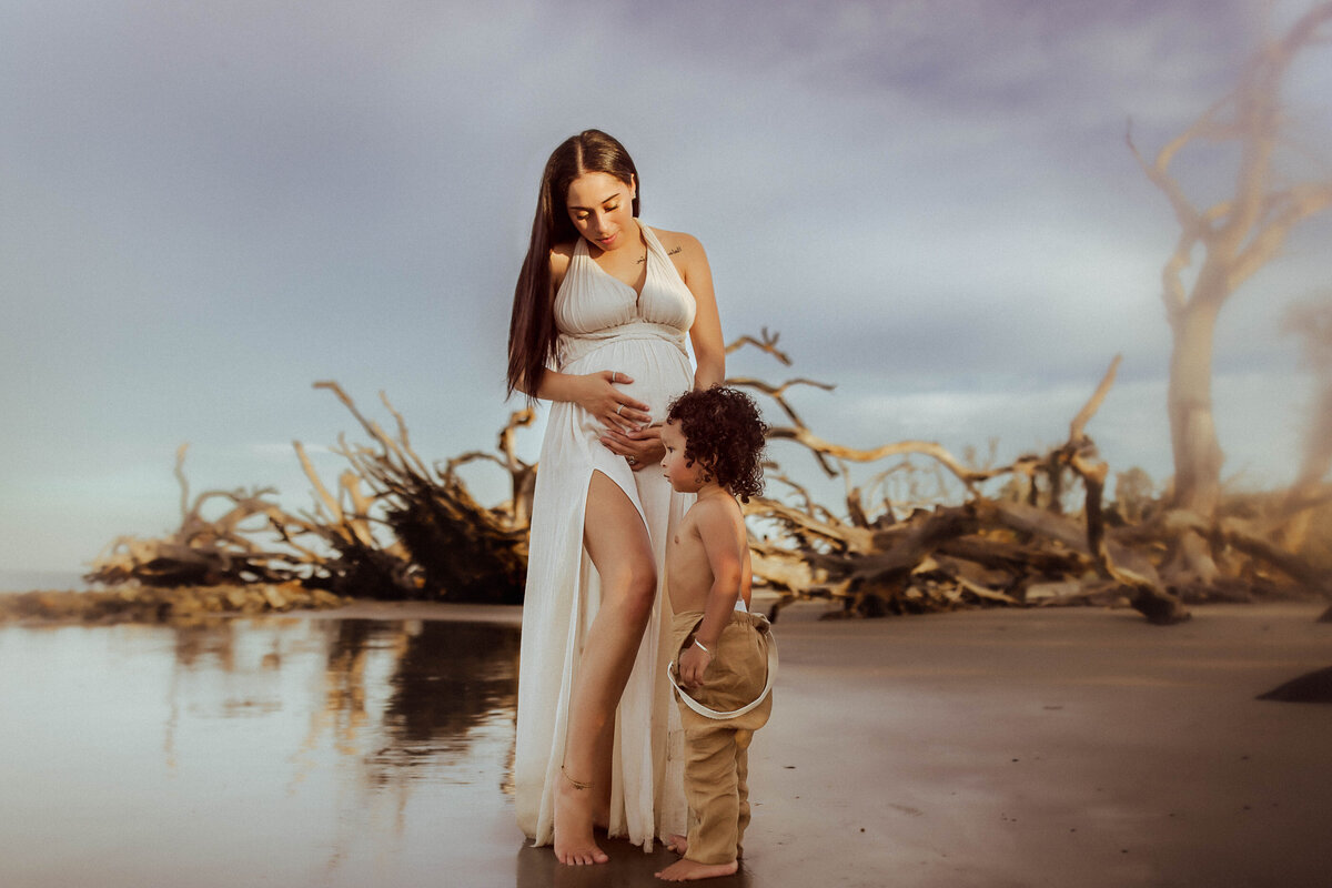 Pregnant woman in a white dress stands by water on a beach with driftwood. A young child in brown pants touches her belly, both gazing down.