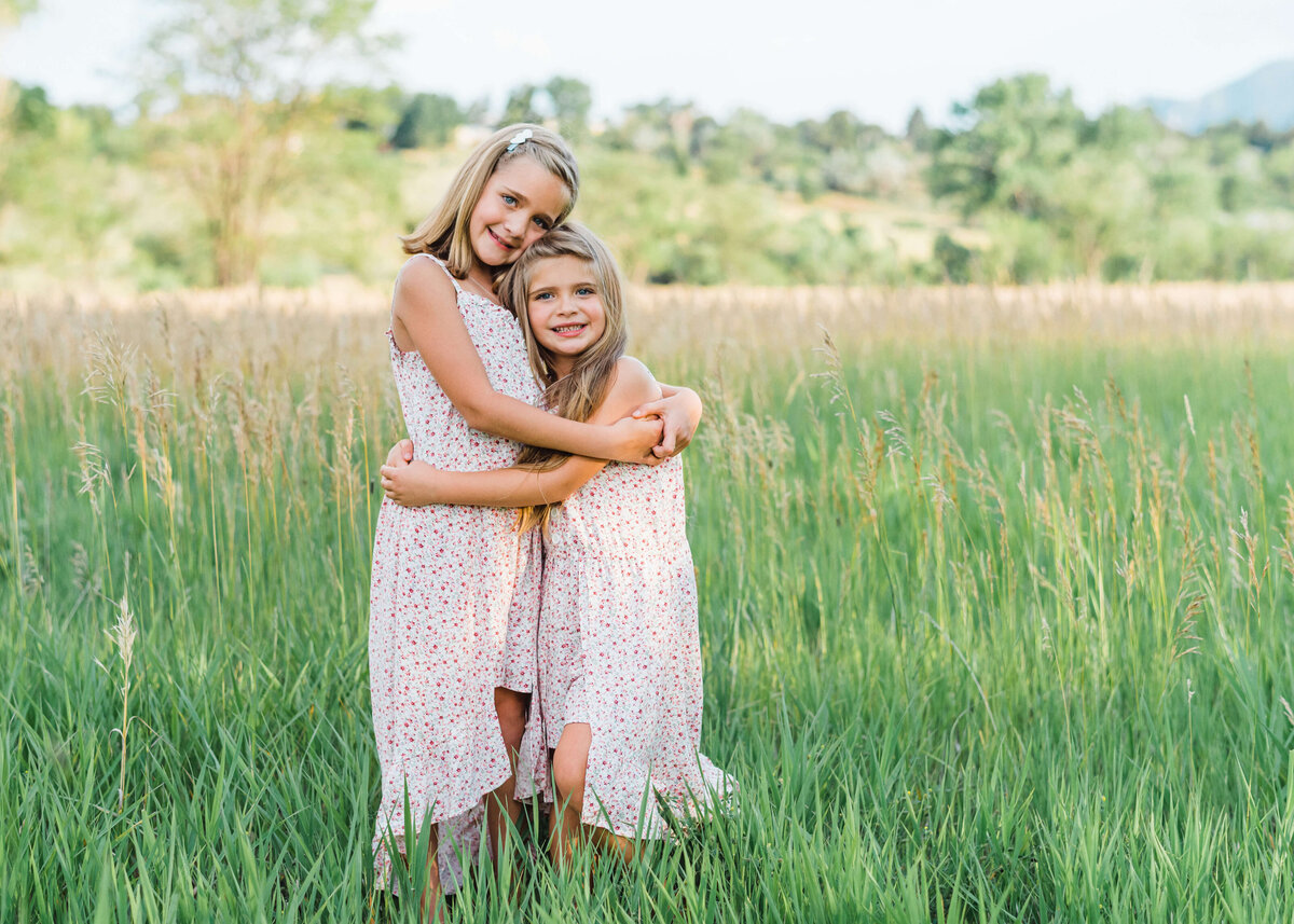 Two blonde girls who are sisters and best friends give each other a hug while standing in a beautiful green field at sunset