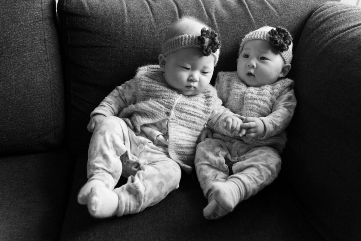 Black-and-white photo of twin baby girls holding hands while sitting together on a sofa, dressed in matching knit vests and headbands.