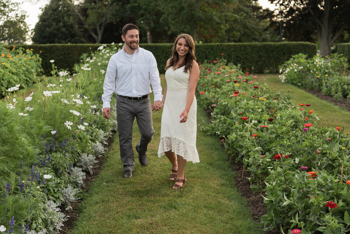 An engaged man and woman are walking, holding hands, down a row of wildflowers in a garden, smiling at the camera
