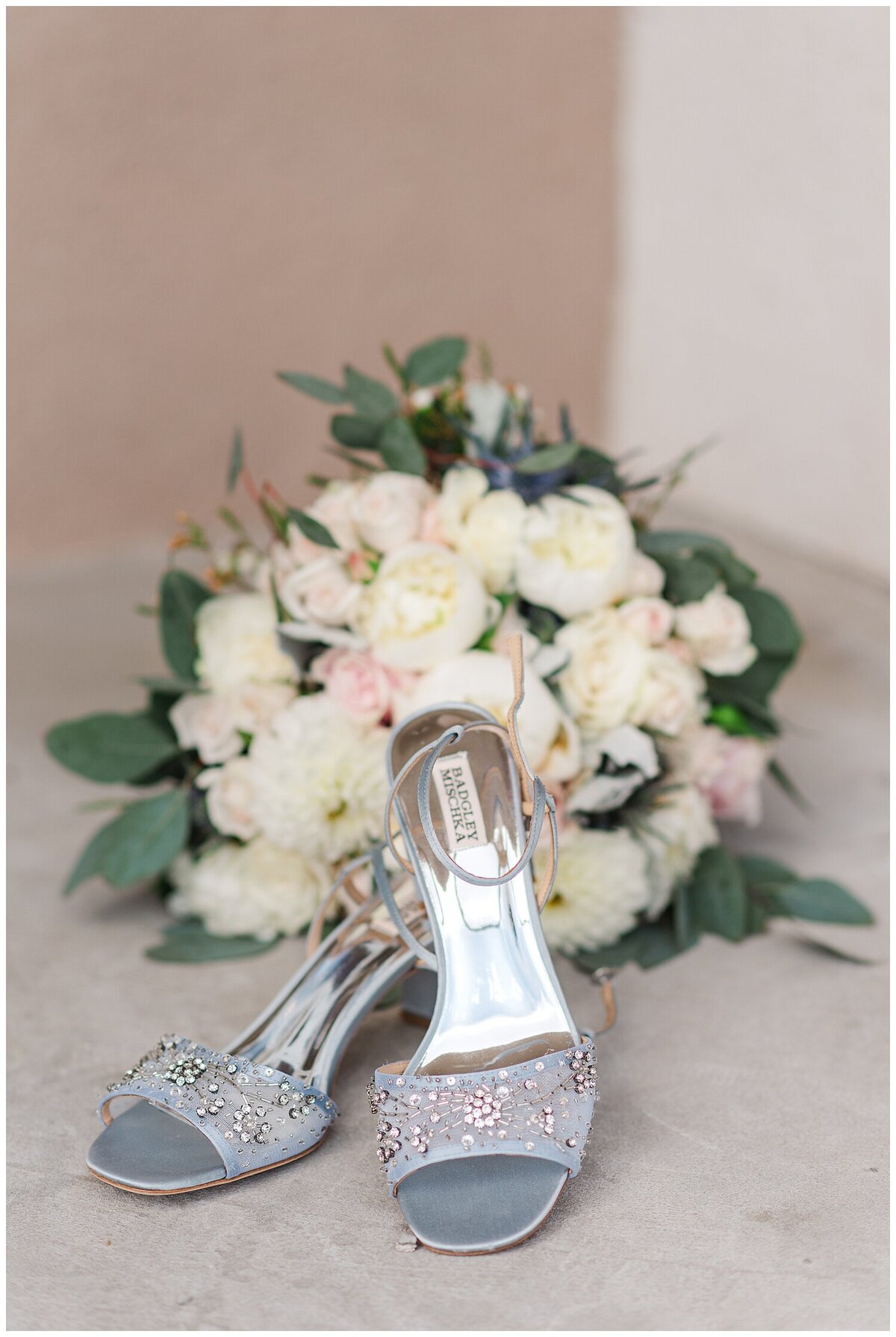 close up photo of a bouquet of white and light pink roses and greenery with dusty blue strap heels in front of it displayed on a concrete floor and a concrete wall in the background