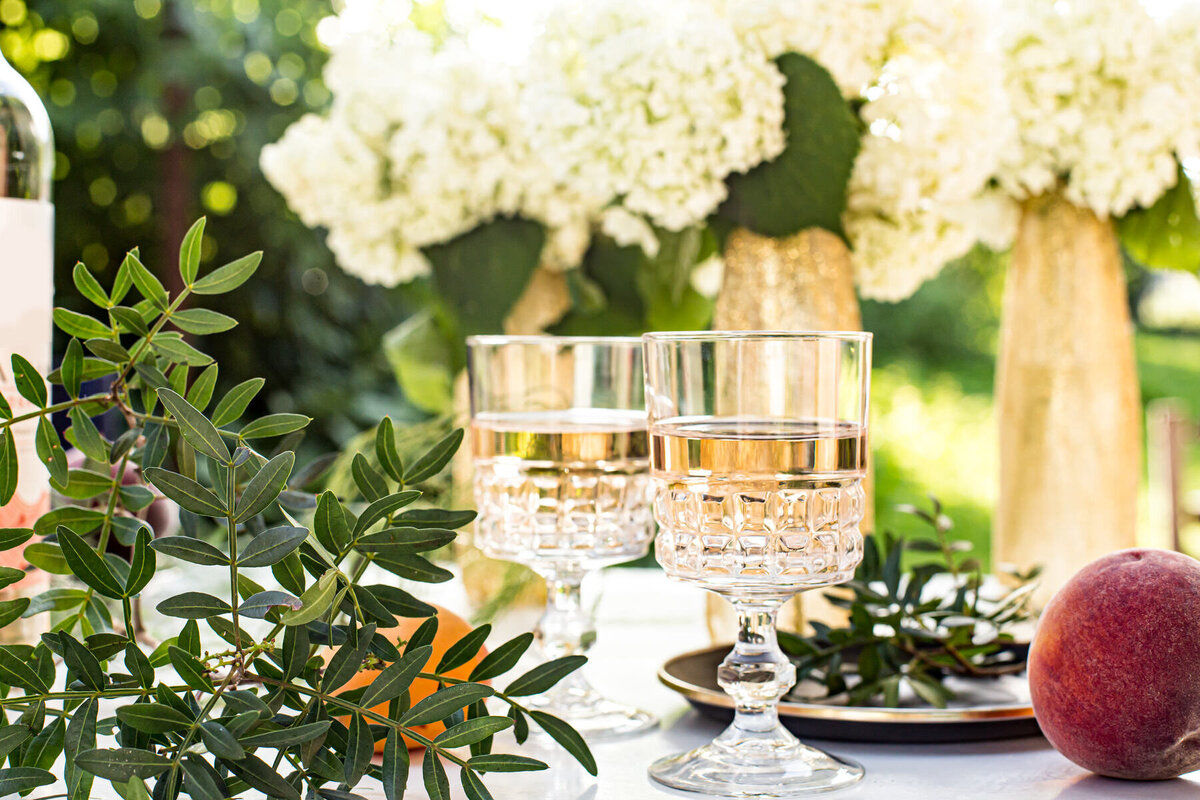 Two crystal glasses filled with rosé wine are set on an outdoor table adorned with green foliage, a peach, and white hydrangea flowers in vases. The scene is sunlit and elegant, suggesting a summer gathering or garden party.