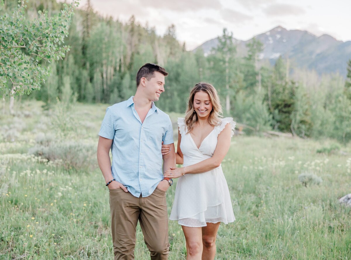 A woman in a white dress holds the arm of her partner while walking in the mountains