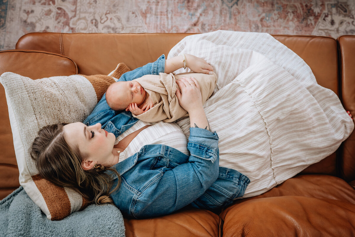 A mother reclines on a leather couch, holding her swaddled newborn close to her chest, both resting comfortably together.