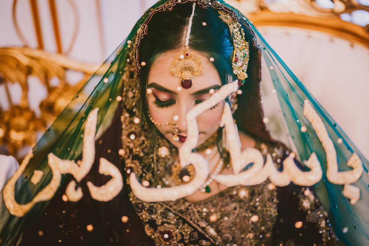 Muslim Pakistani bride looking down at her Nikkah ceremony with an arabic print dupatta veil over her head