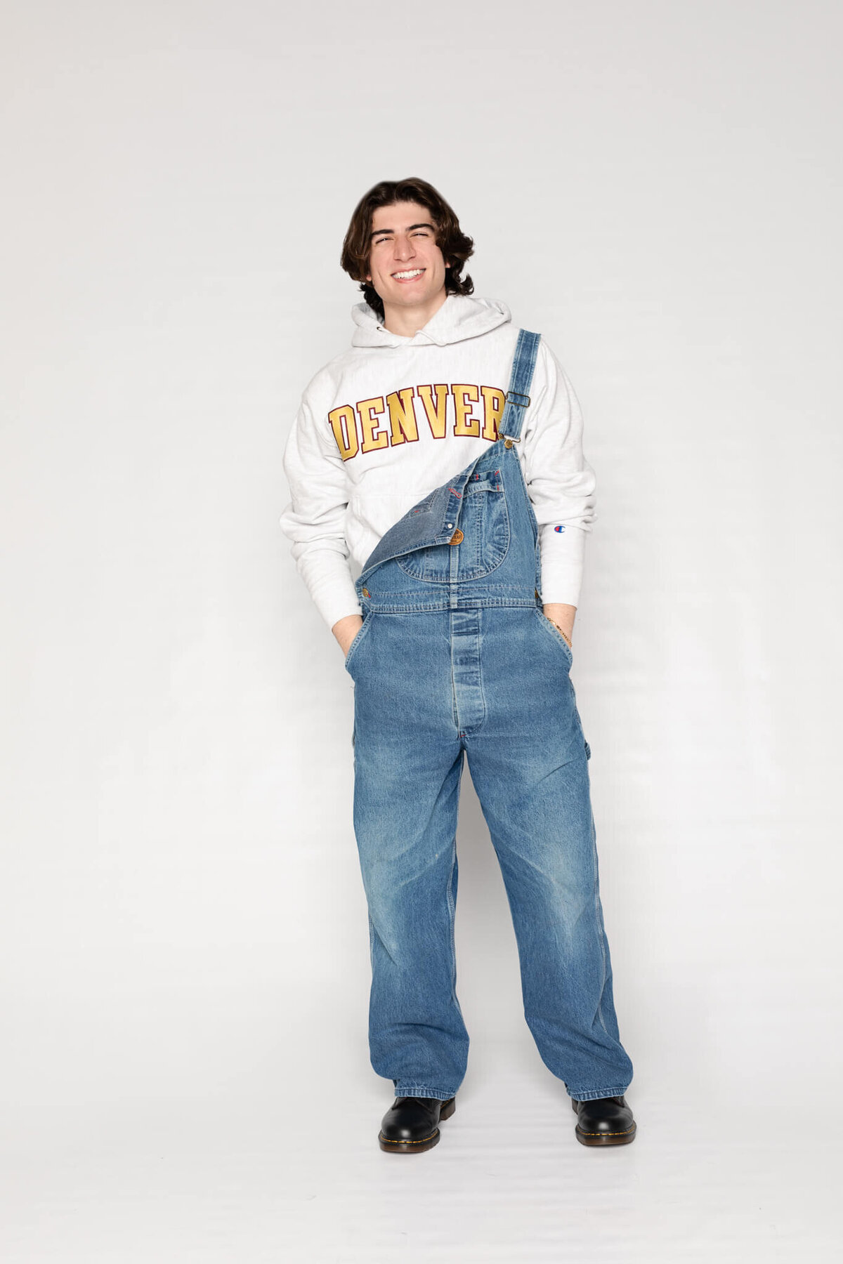 A person wearing a white Denver hoodie under blue denim overalls stands smiling with hands in pockets against a plain background. They have dark hair and are wearing black shoes.