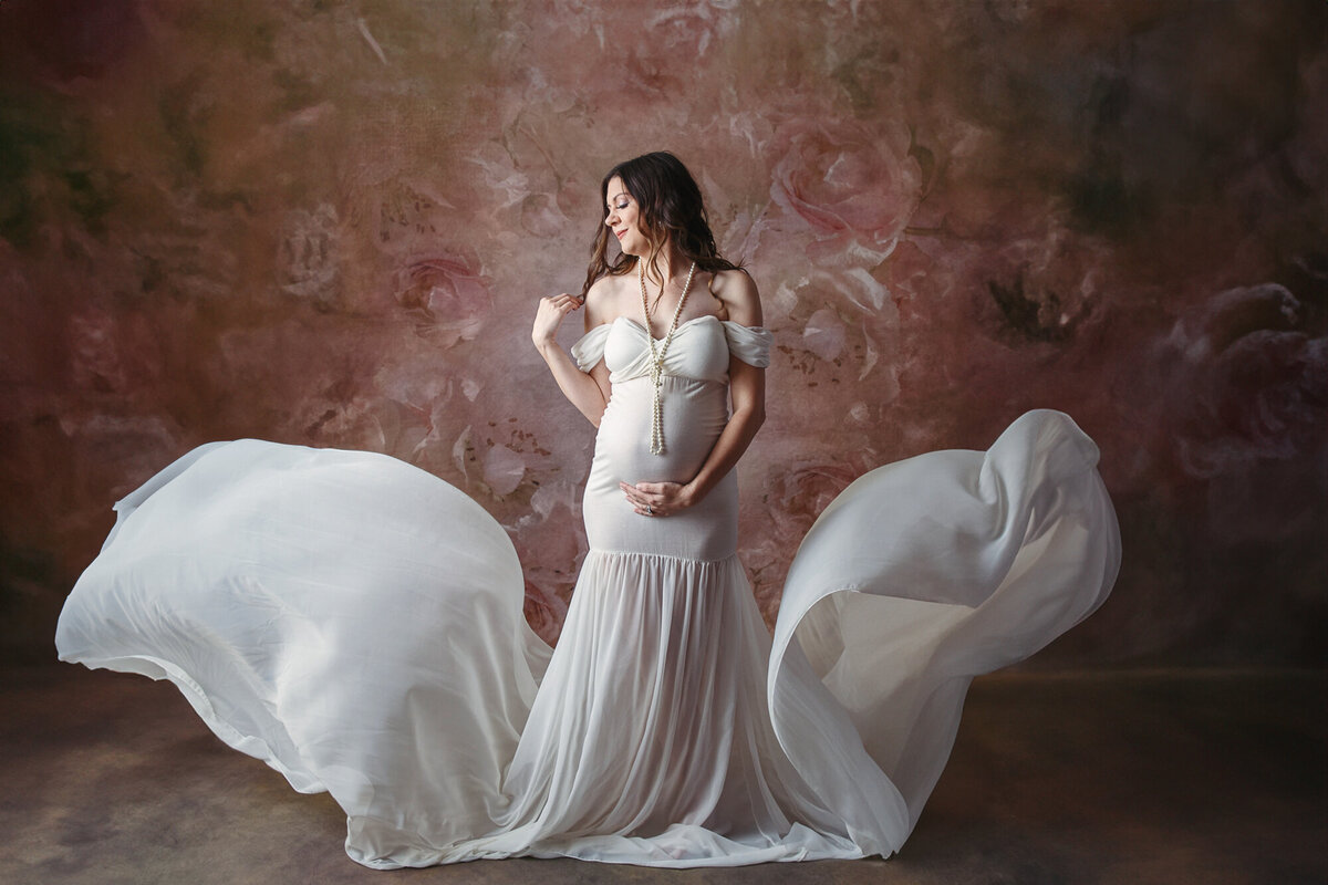 Beautiful portrait of a pregnant woman wearing a white gown and white pearls photographed on a brown floral studio background