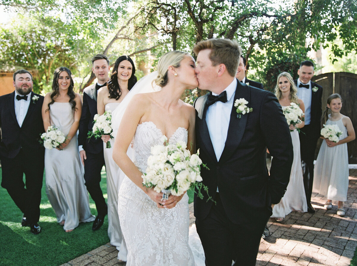 A stunning bride and groom share a romantic kiss as they walk hand in hand, surrounded by joyful friends and family. Captured in natural light, this fine art wedding photograph highlights the love and celebration of a beautiful wedding day. Photographed at an elegant venue with lush greenery in the background.