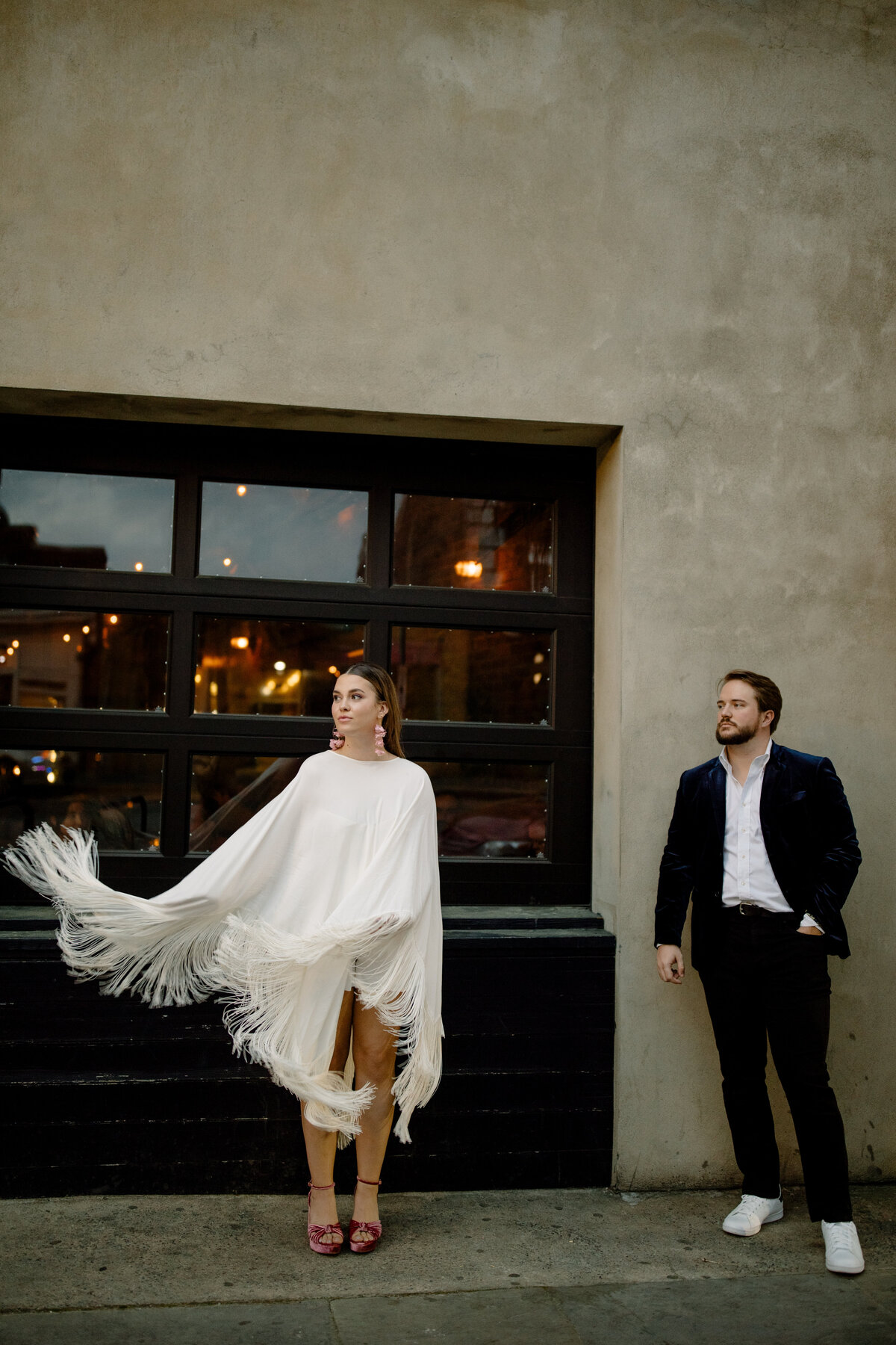 Bride in fringe poncho standing in front of garage door with groom admiring