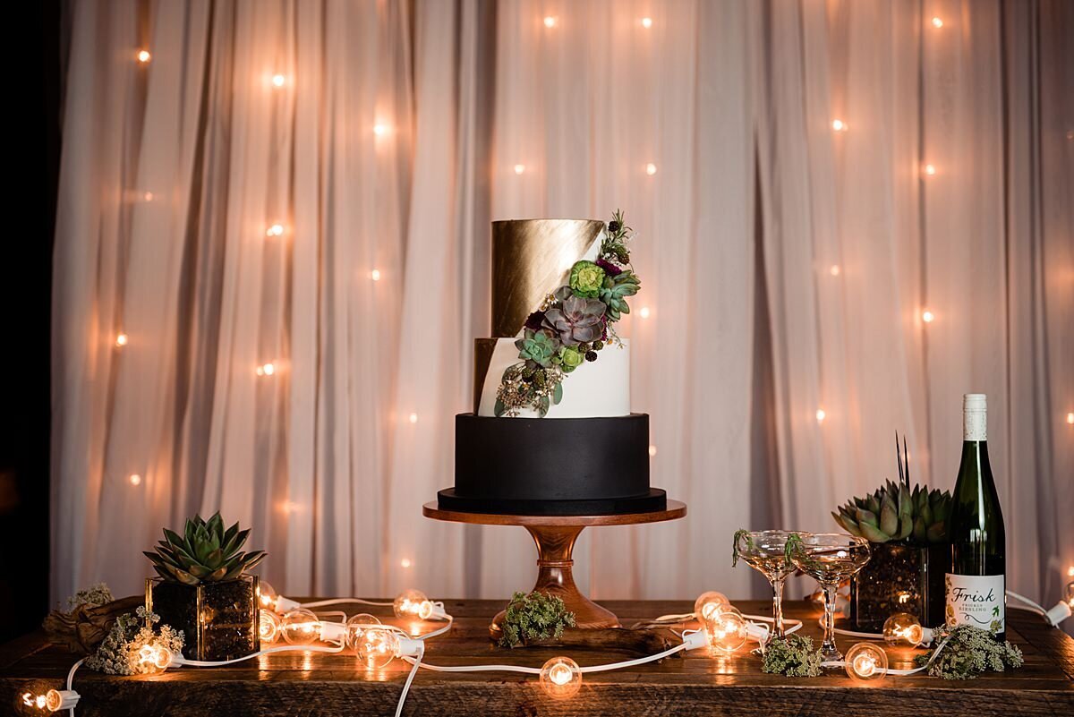 Against a wall covered with sheer white drapery and string lighting, sits a farm table with a black, white and gold three tier wedding cake on a footed wood stand. The cake is decorated with succulents, black raspberries and brussel sprouts. A bottle of wine sits on a table next to a small planter with a succulent and a strand of string lighting at City Winery Nashville.