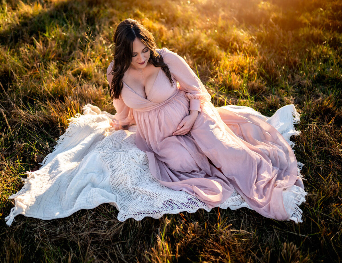 A beautiful mama to be sits on a blanket in the grass and cradles her baby bump during her portrait session with an Asheville Maternity Photographer