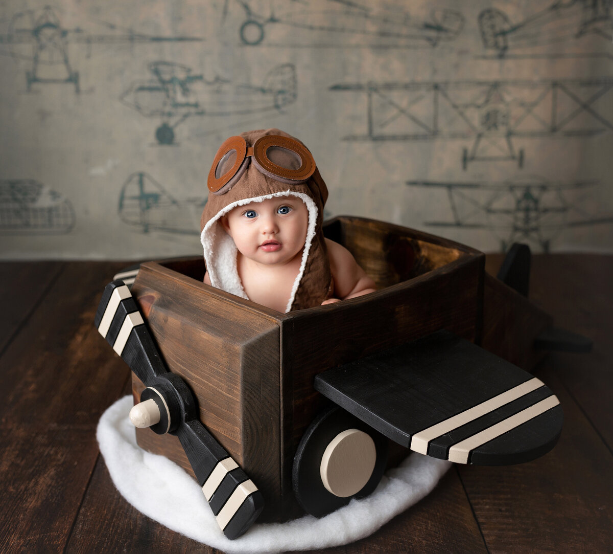 Baby boy 6-month milestone photo shoot in West Palm Beach and Boynton Beach photography studio.  Baby boy is sitting in an airplane inspired crate and vintage pilot cap. Baby is looking at the camera. Backdrop is vintage plane sketches.