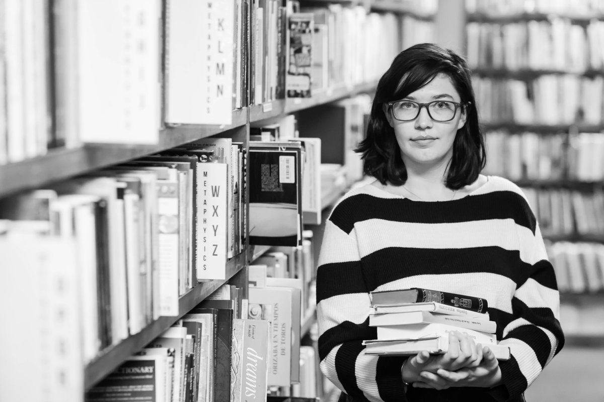 black and white photo taken at Books in stock bookstore wooster ohio, high school girl with dark hair wearing black and white stripped sweater holding a stack of books standing in an aisle of books, photographed using the thirds rule so books are closer than client with lots of bokah, photographed by Jamie Lynette Photography Canton Senior Photographer