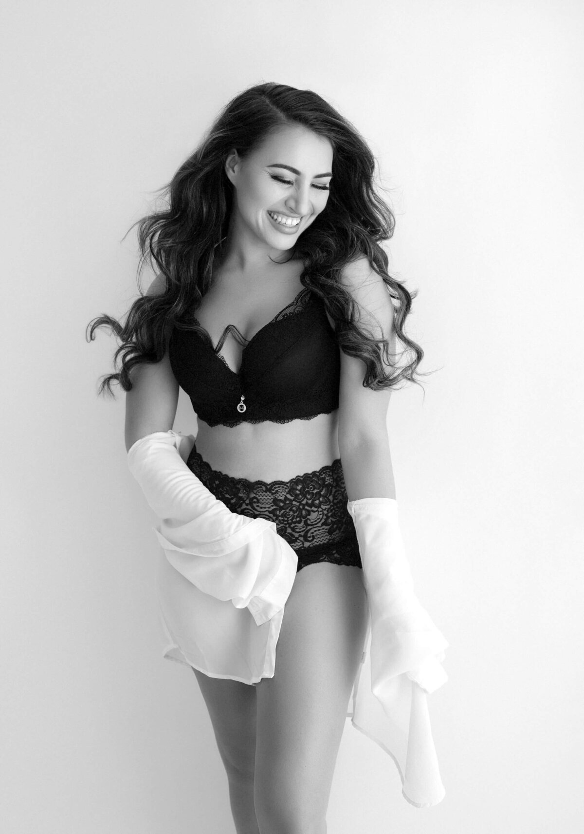 A joyful woman in black lace lingerie and sheer white sleeves laughs with radiant confidence, captured in a bright and airy boudoir setting.