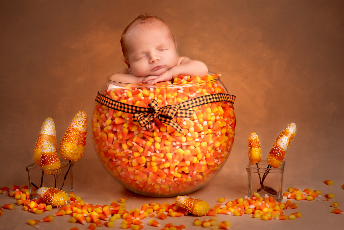 Newborn nestled in a bowl of candy corn with warm fall decor, captured in Overland Park, Kansas. Perfect for families looking for autumn-themed newborn photography in the Kansas City area