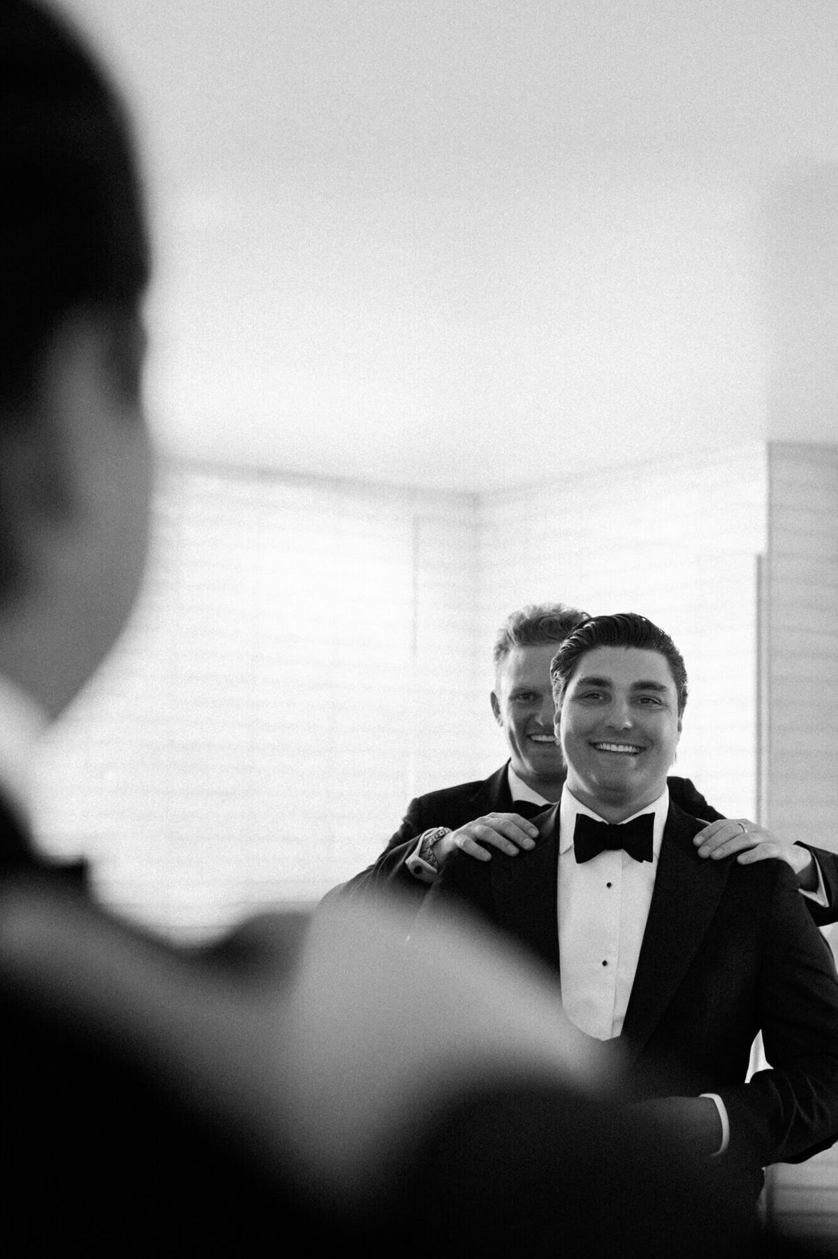 Groom smiling in the mirror as his best man adjusts his suit jacket before the ceremony, captured by a luxury wedding photographer in Scottsdale.
