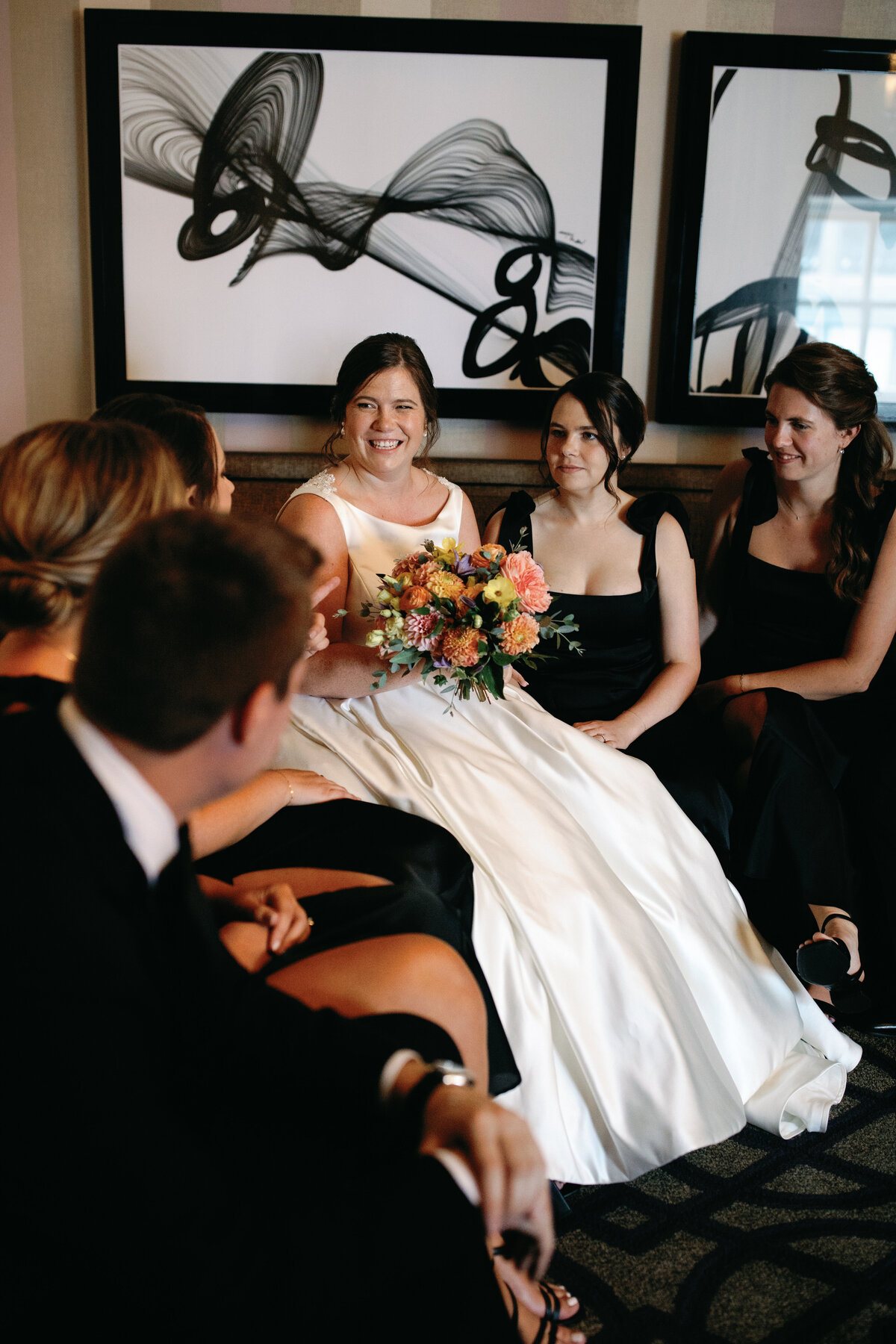Bride in satin ballgown sitting with bridesmaids in black dresses, laughing together at Hotel Monaco Washington DC.