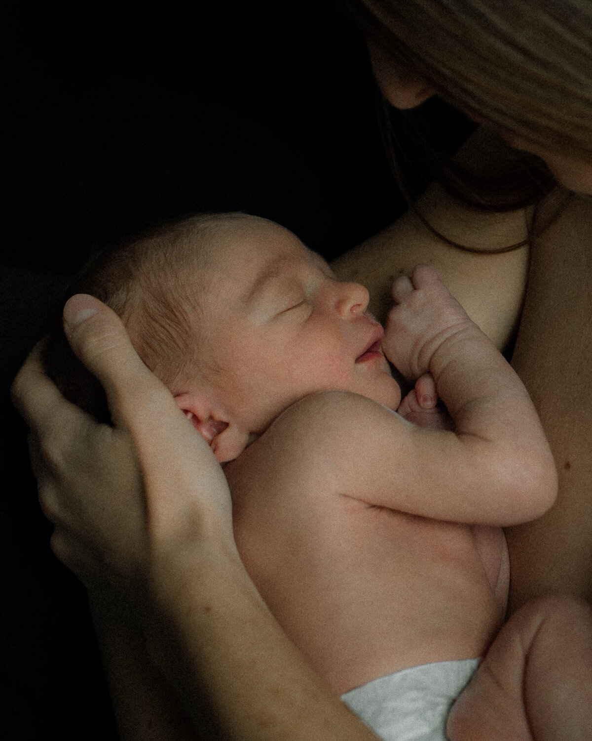 Close-up of newborn baby resting on mother’s chest in soft, moody light.