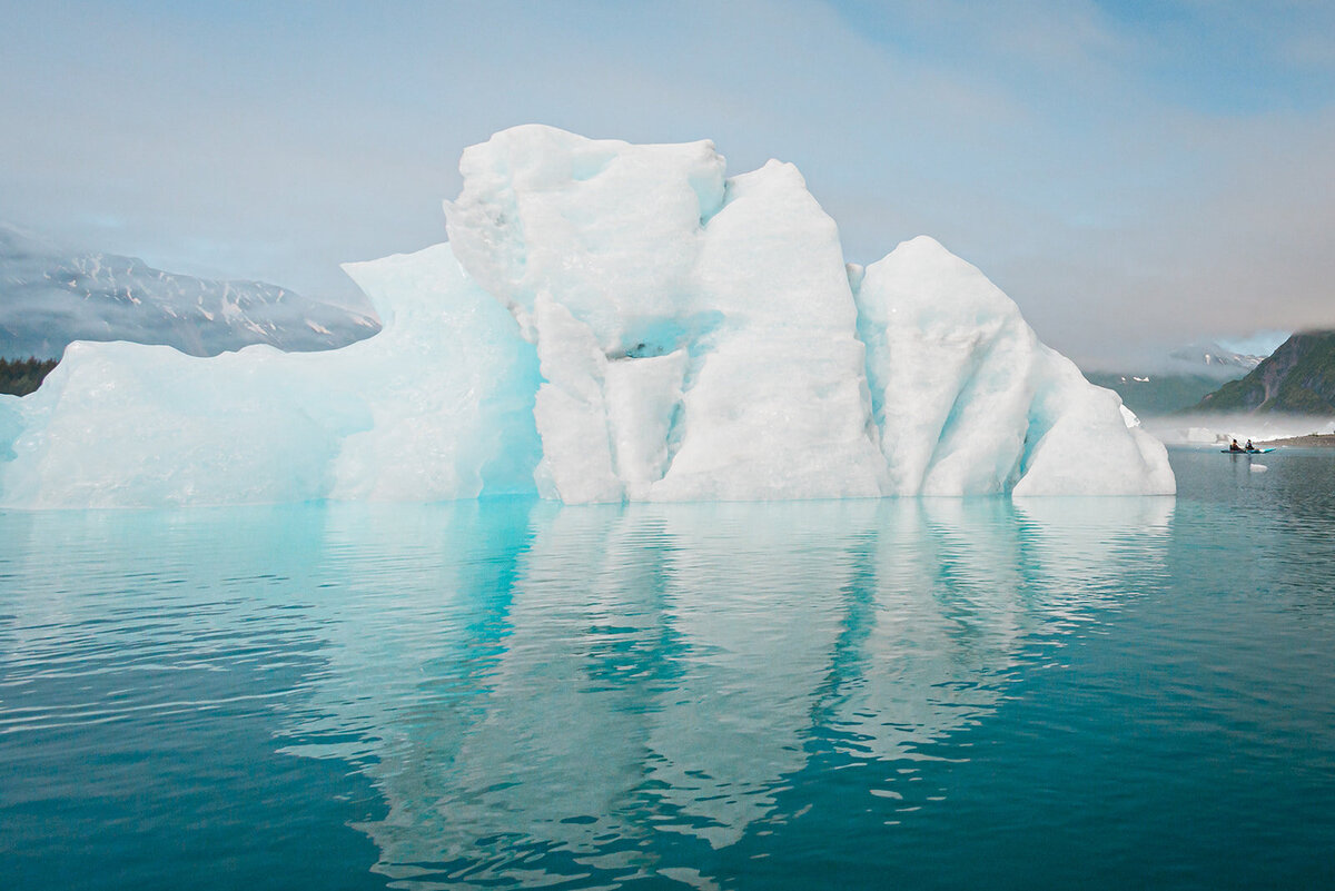 Filling the view is a large blue-tinted white iceberg shaped like an arrow and it's reflecting, floating in the beautiful turquoise water of Bear Lake during a two day luxury elopement with dogsledding in Alaska.  On the right side of the iceberg at the "arrow" tip, is a tiny kayak with the bride and groom in it as they explore during their elopement.